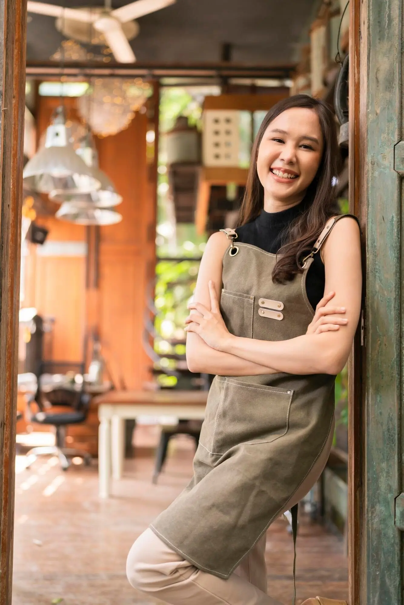 Retrato de una mujer adulta asiática con delantal, de pie en la entrada de su estudio-taller de cerámica en casa, vestida de manera informal, relajada, sonriente, segura y acogedora.